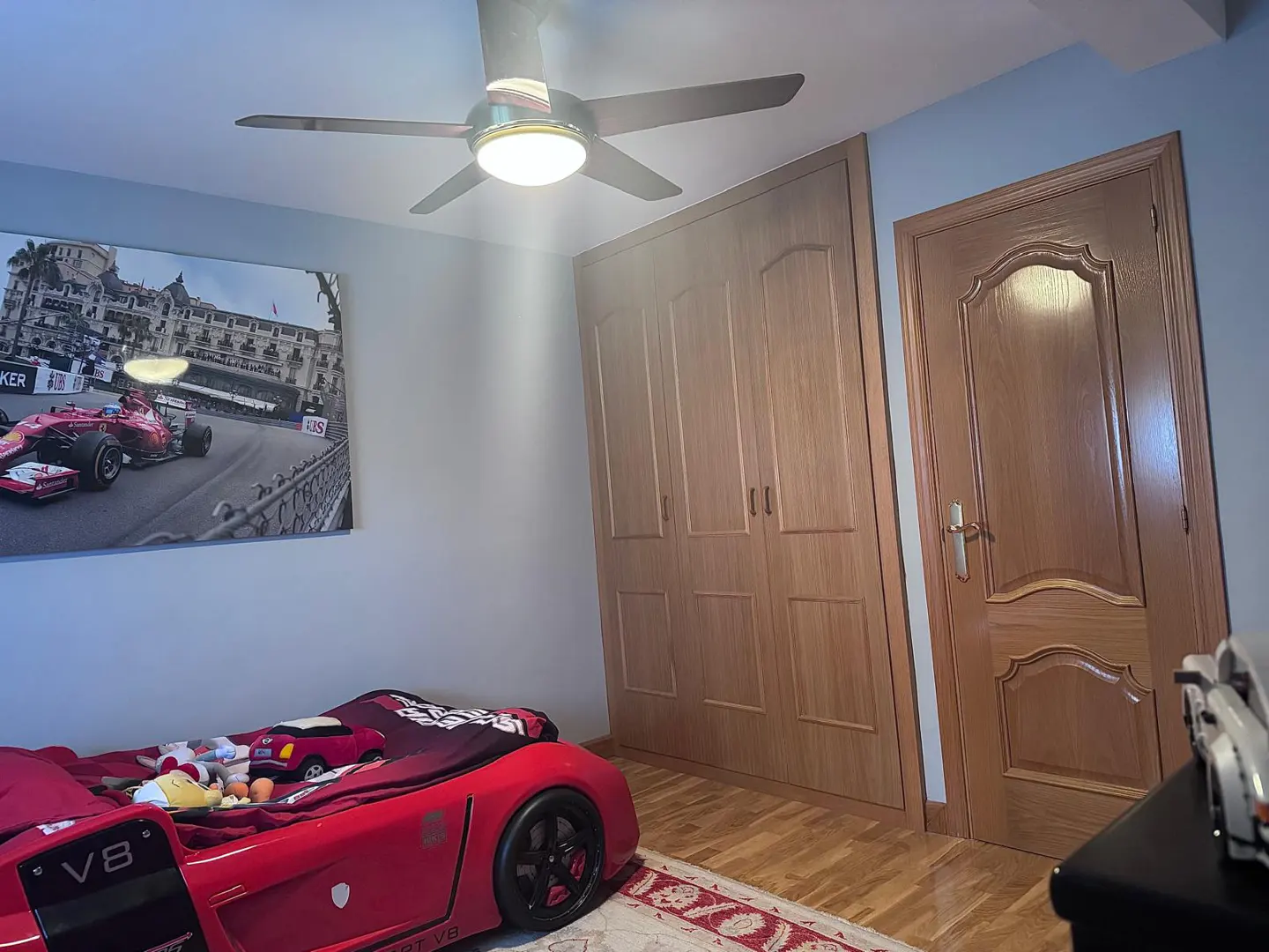 A child's bedroom with a red race car bed, a ceiling fan, and a large racing poster on the wall. The room has light blue walls and a wooden floor.