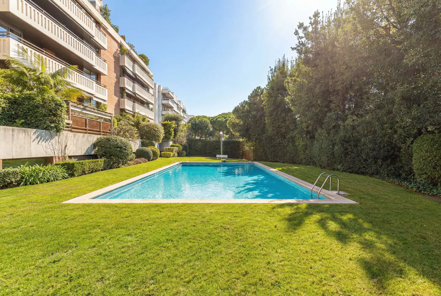 A rectangular swimming pool with blue water is surrounded by green grass and trees. Apartment buildings are in the background.