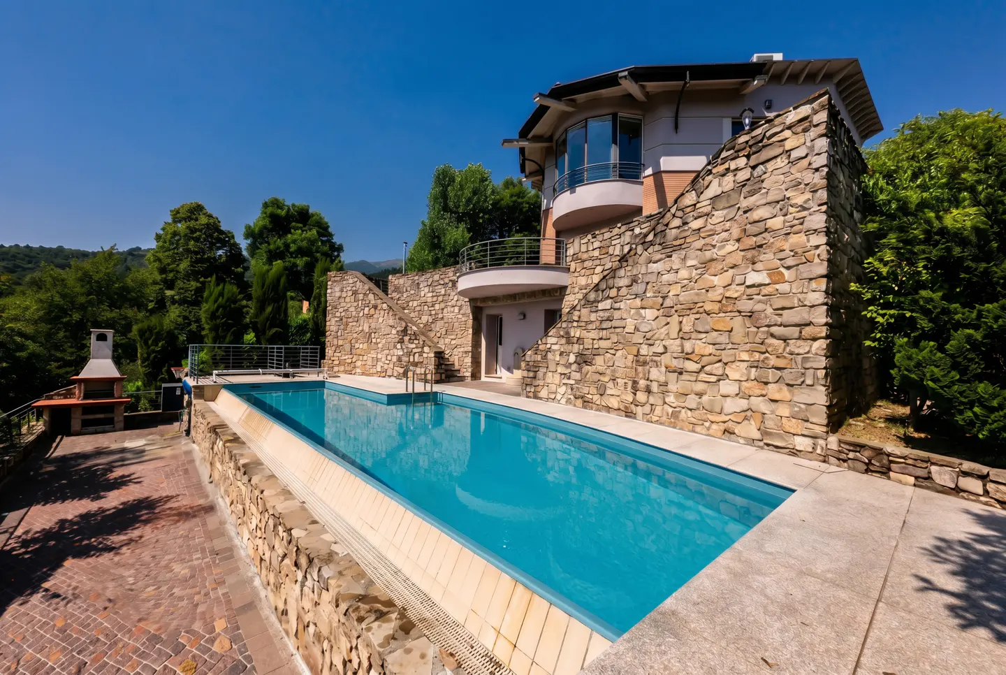 Modern home with a stone facade and balcony overlooking a rectangular swimming pool on a sunny day.