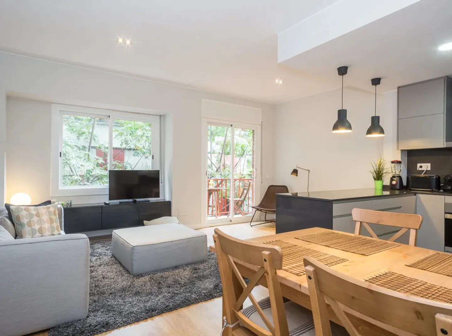 Modern open-plan living area with a dining table, grey sofa, TV, and a kitchen with dark grey cabinets and pendant lights.