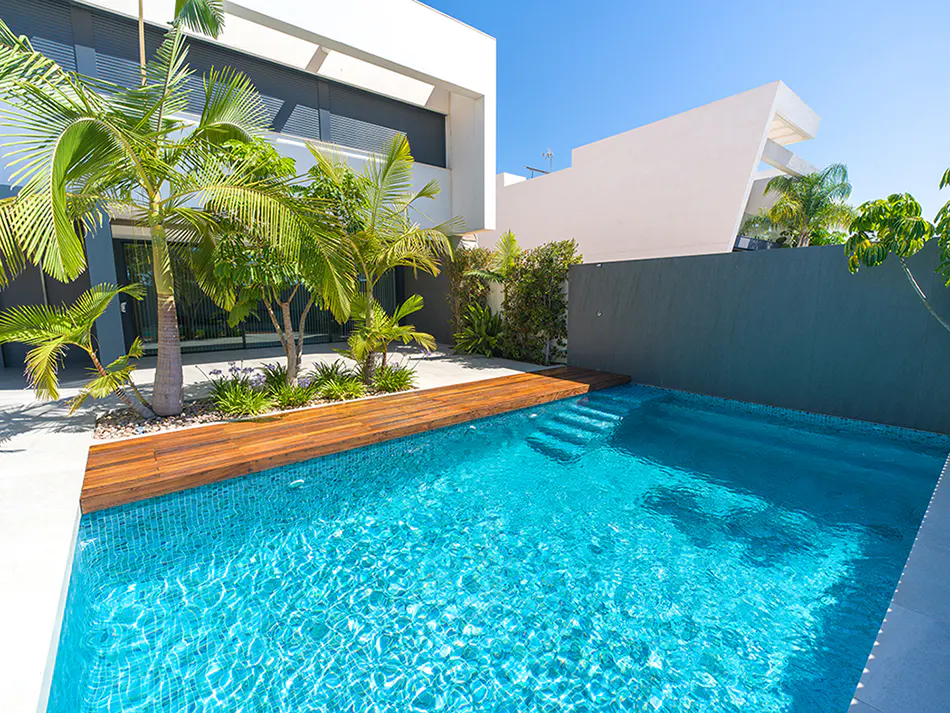 Modern home with a bright blue swimming pool, wooden deck, and palm trees against a clear blue sky.