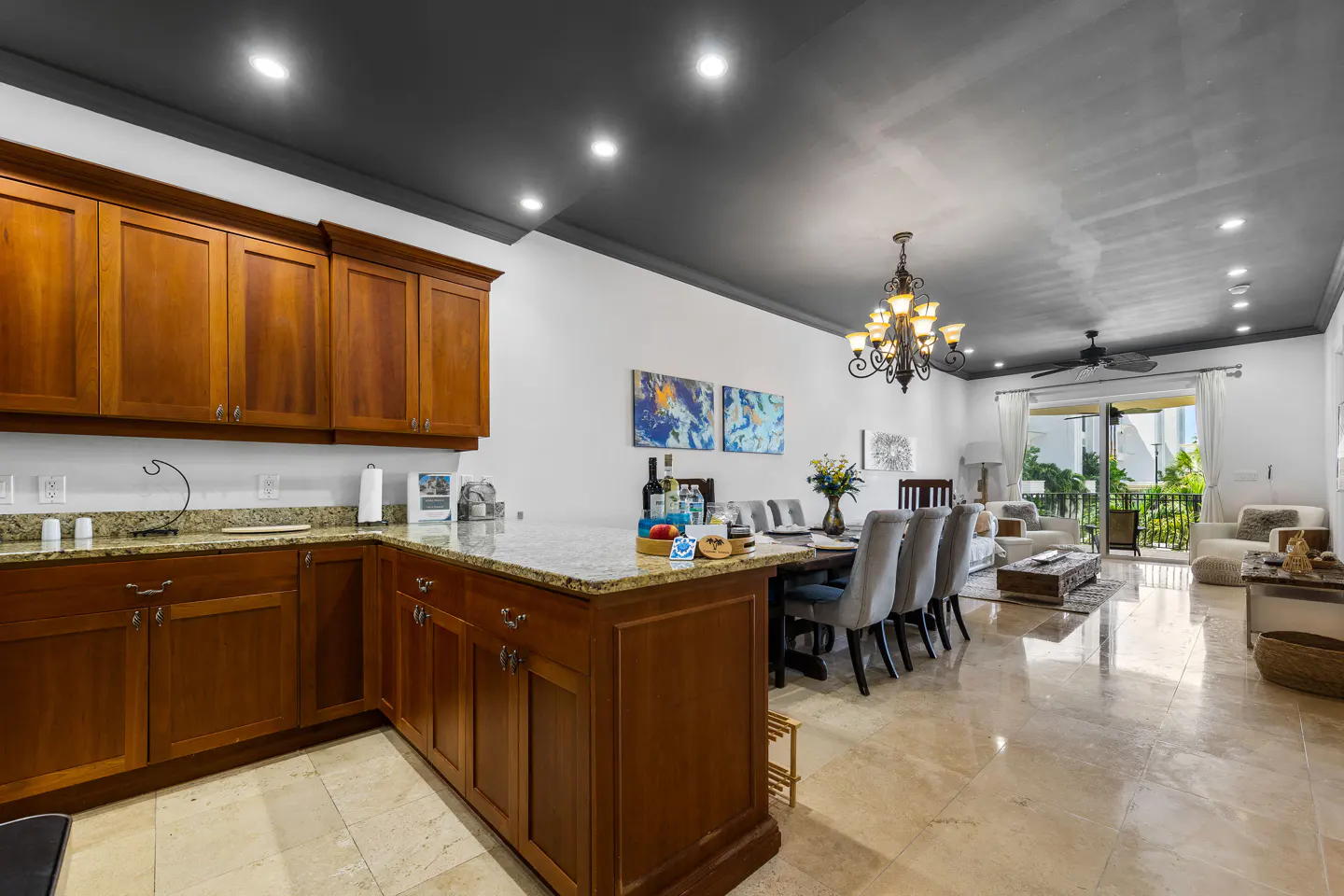 Open-concept kitchen, dining, and living area with brown cabinets, granite countertops, and marble floors. A chandelier hangs above the dining table.