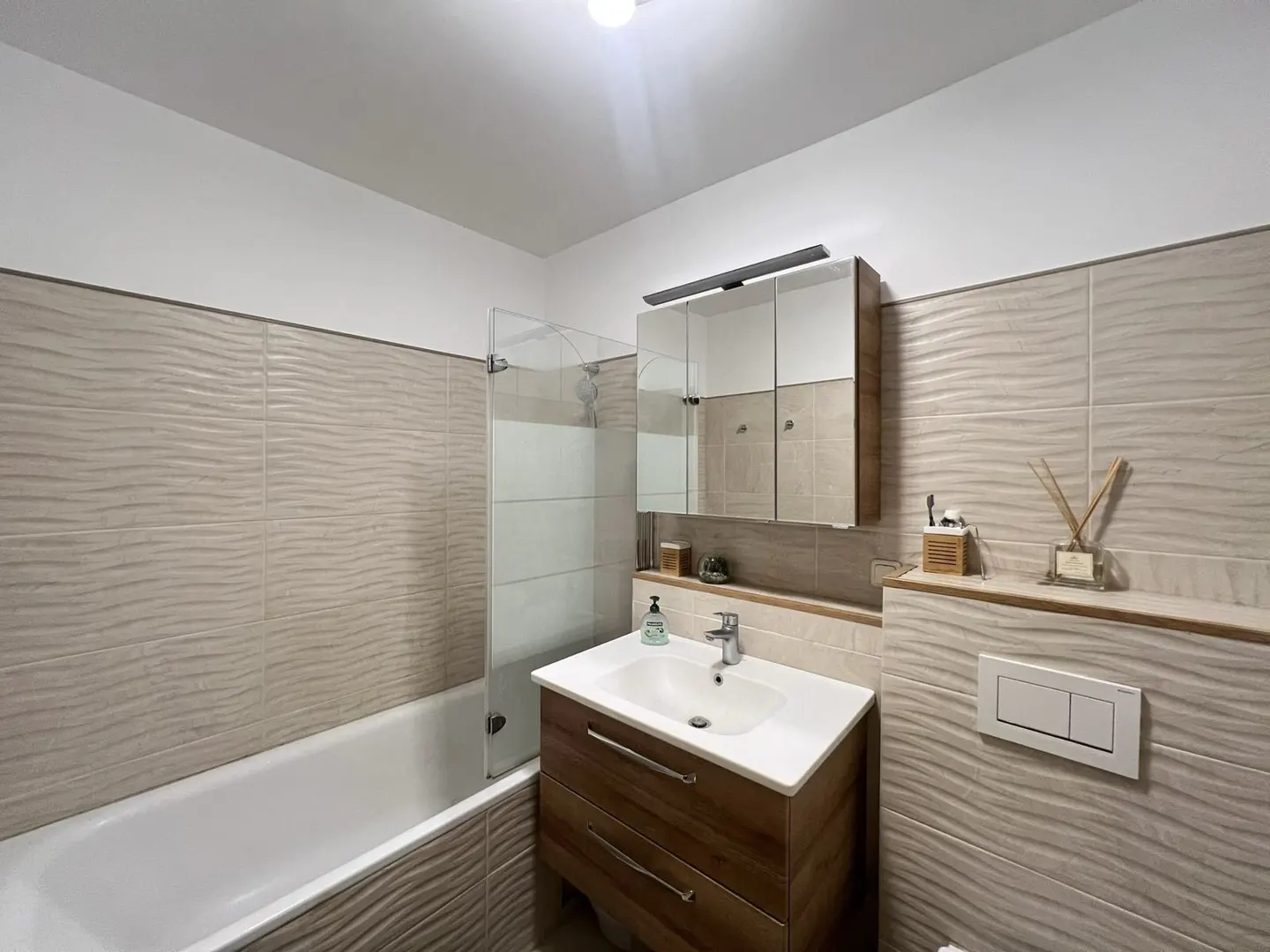 Bathroom with textured beige tiles, white tub with glass door, wood vanity, and mirrored cabinet.