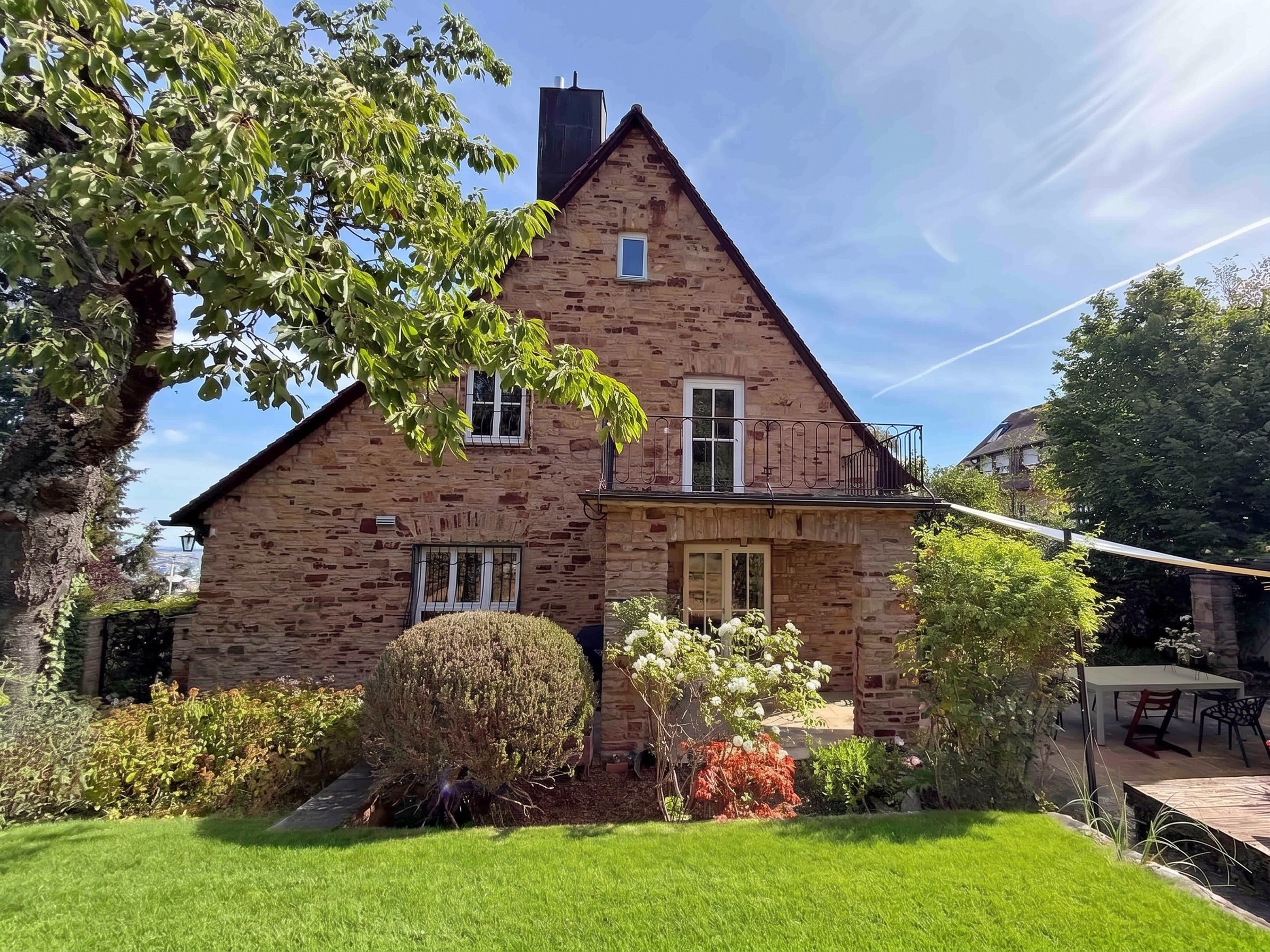 A rustic stone house with a balcony, surrounded by a lush green lawn, trees, and a patio area under a bright blue sky.