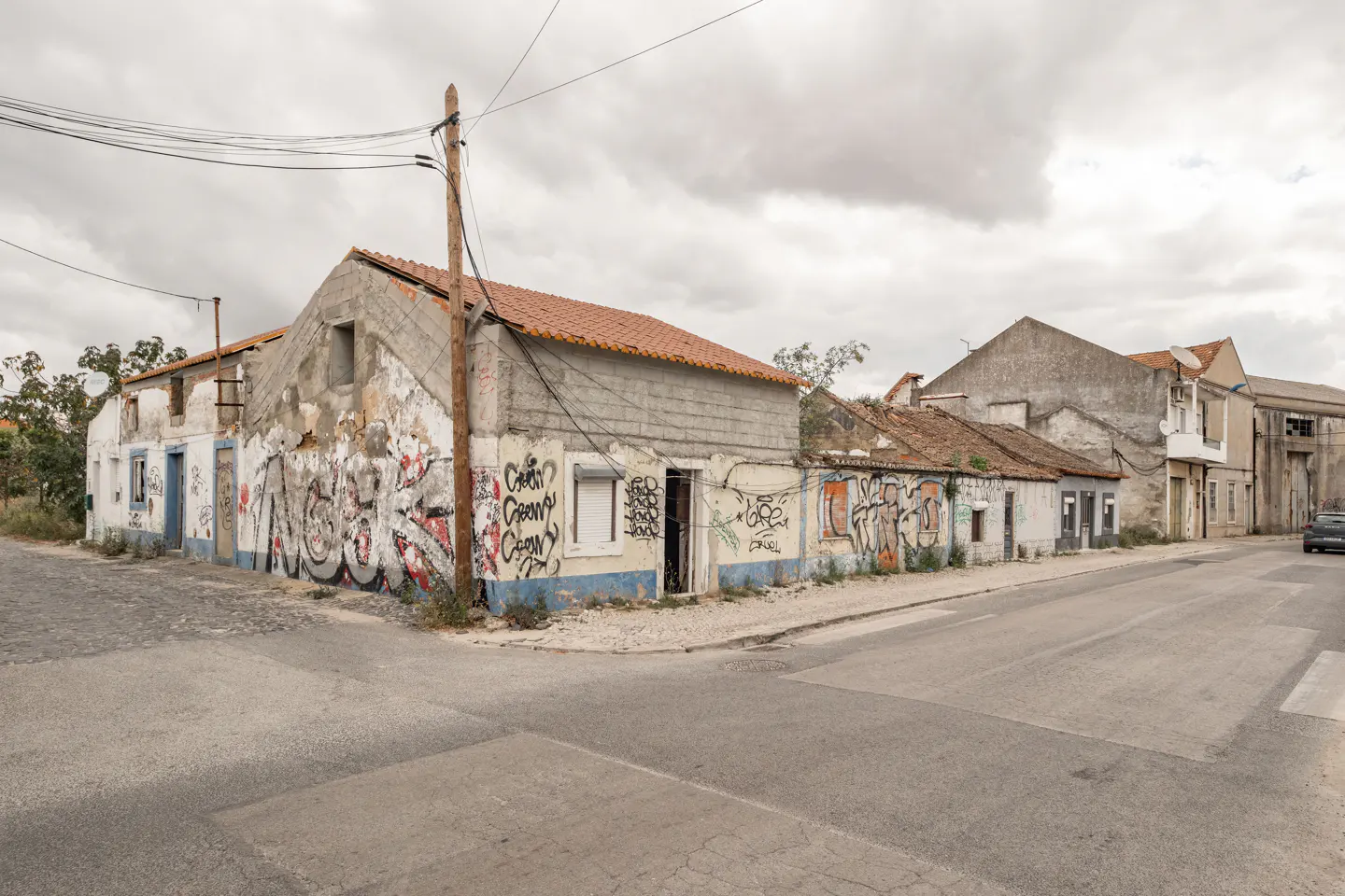 Street view of dilapidated, graffiti-covered buildings with red tile roofs under a cloudy sky. A utility pole stands on the corner.