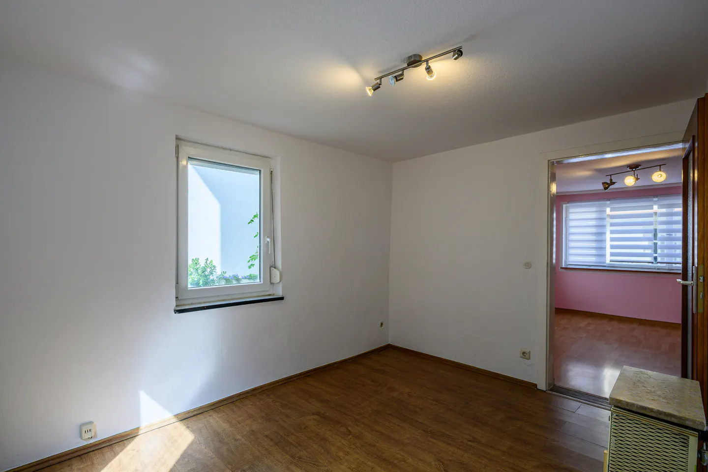 Bright, empty room with wood floor, white walls, and a window. A doorway leads to a pink room with blinds. Track lighting on the ceiling.