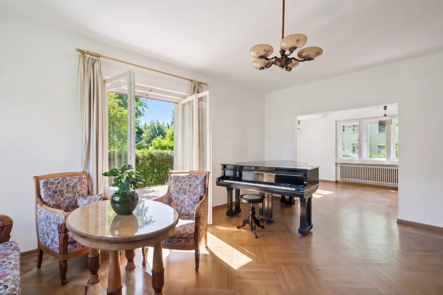 Bright living room with a black grand piano, a round wooden table, floral armchairs, and an open door to a garden.