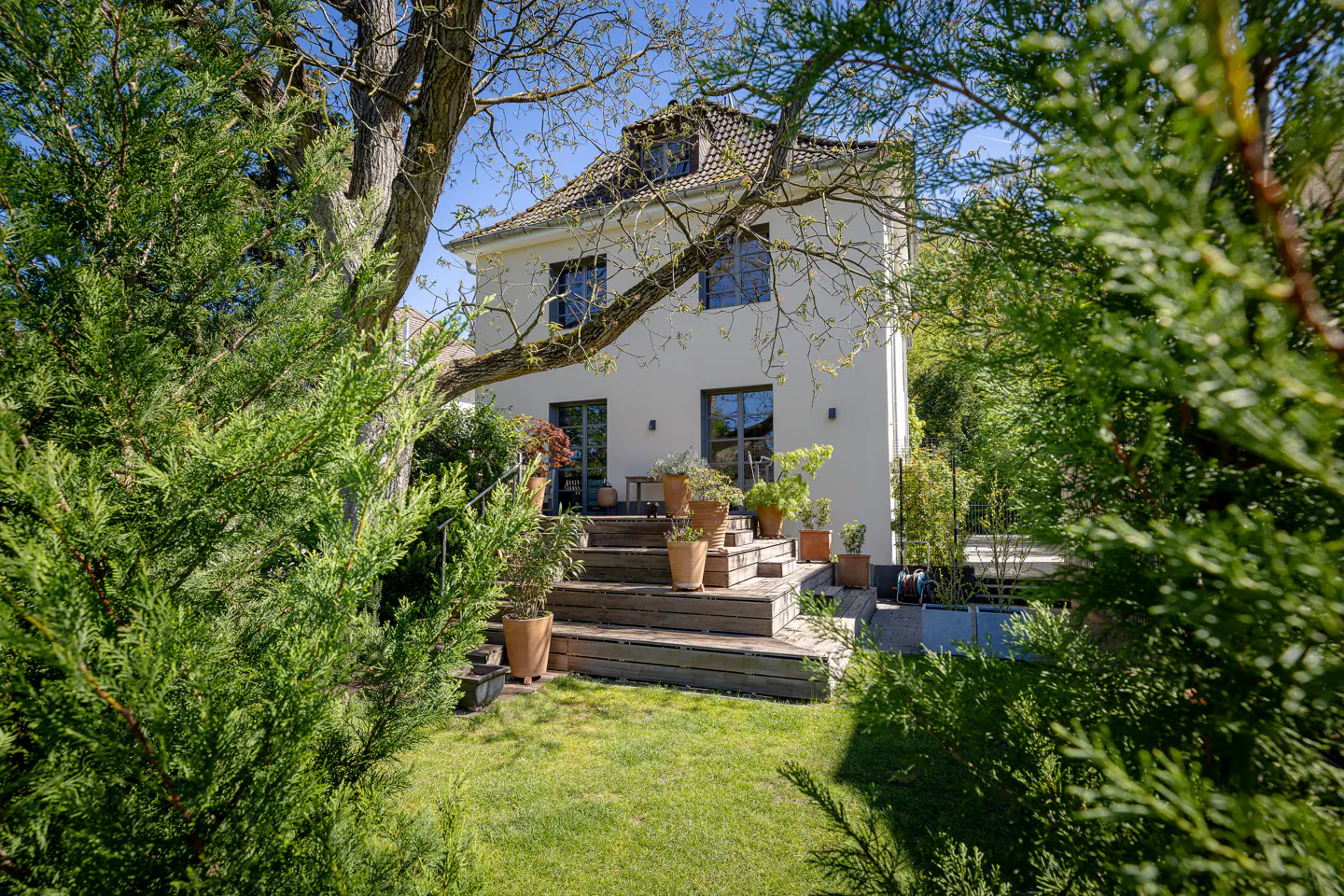 A white two-story house with a brown roof, surrounded by green trees and a lawn. A wooden staircase leads to a patio with potted plants.
