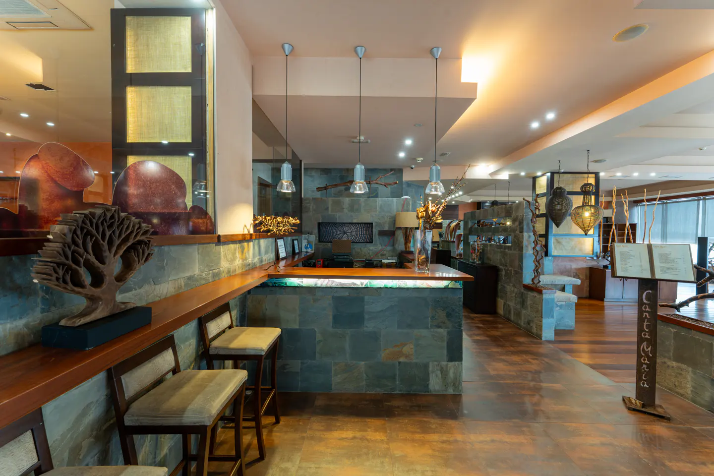 Hotel lobby with stone reception desk, wood floors, and pendant lights. A wooden tree sculpture sits on the counter.