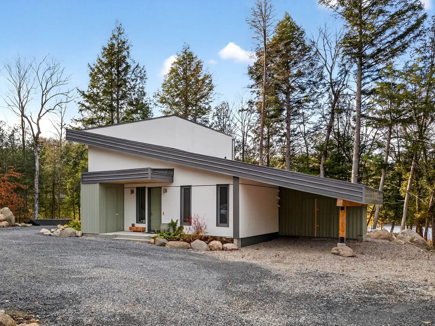 Modern white house with a slanted gray roof and green trim, surrounded by tall trees and a gravel driveway.