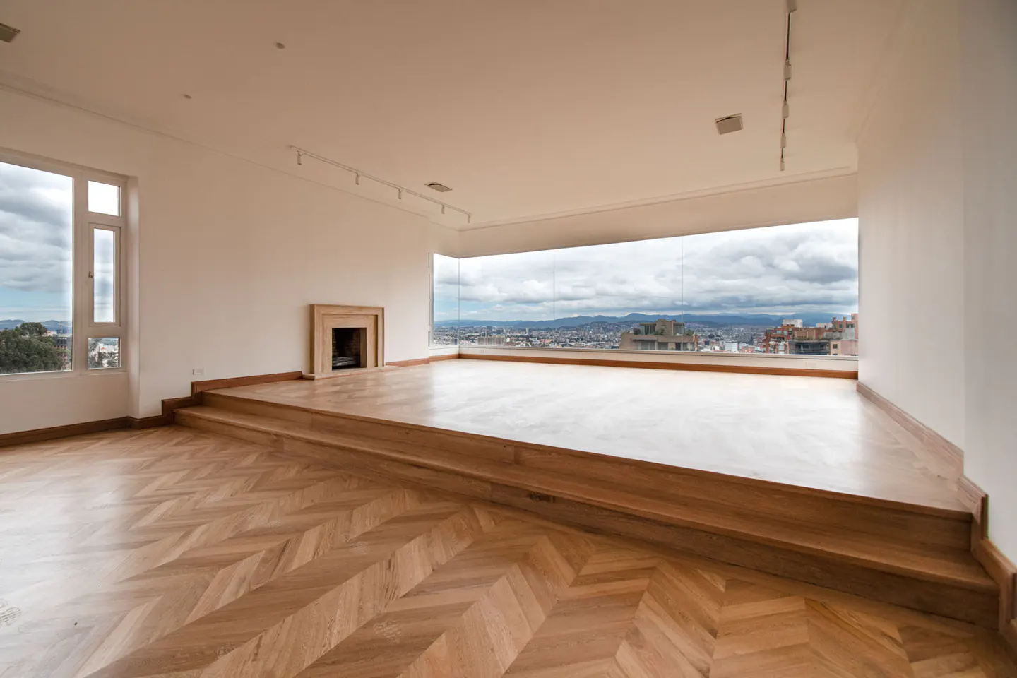 Bright, empty room with herringbone wood floors, fireplace, and large window overlooking a city.