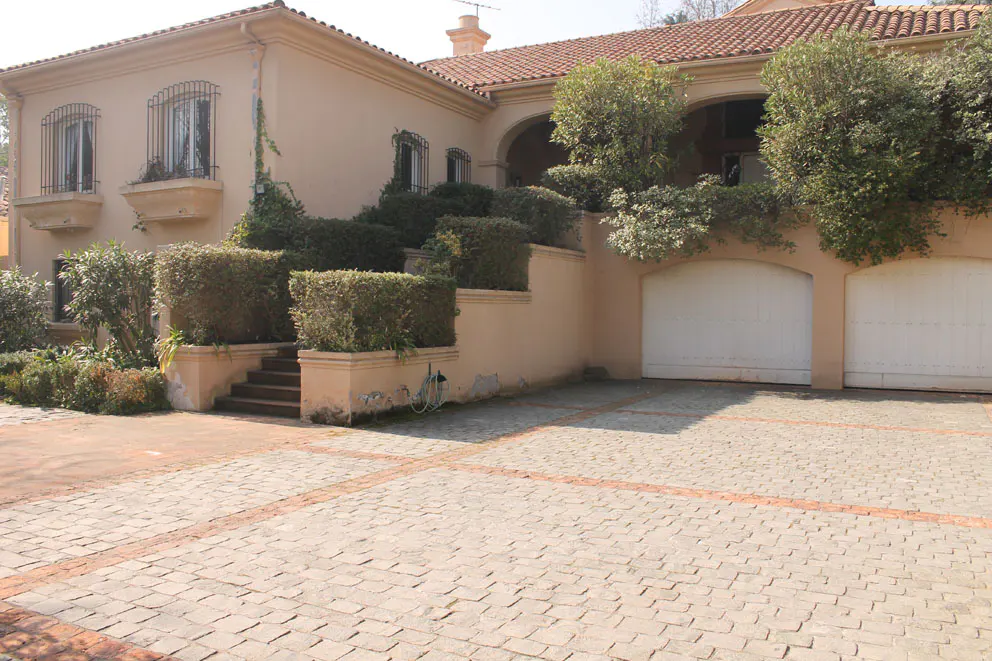 Exterior of a tan stucco house with a red tile roof, manicured hedges, and a cobblestone driveway.