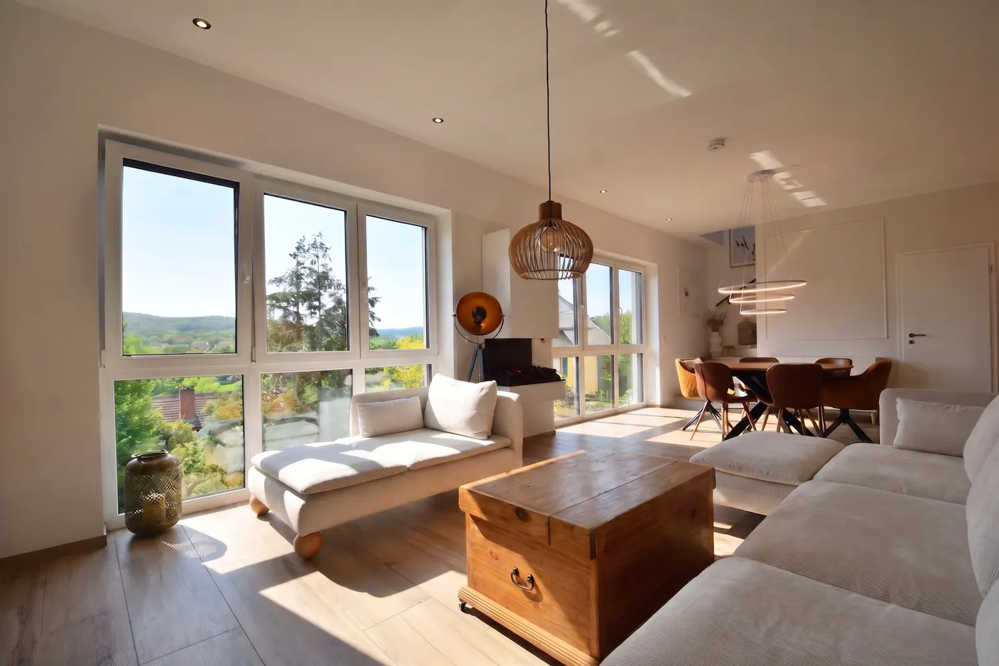 Bright, modern living room with a large window view. A beige sofa, wooden chest, and dining table are visible. Sunlight streams in.