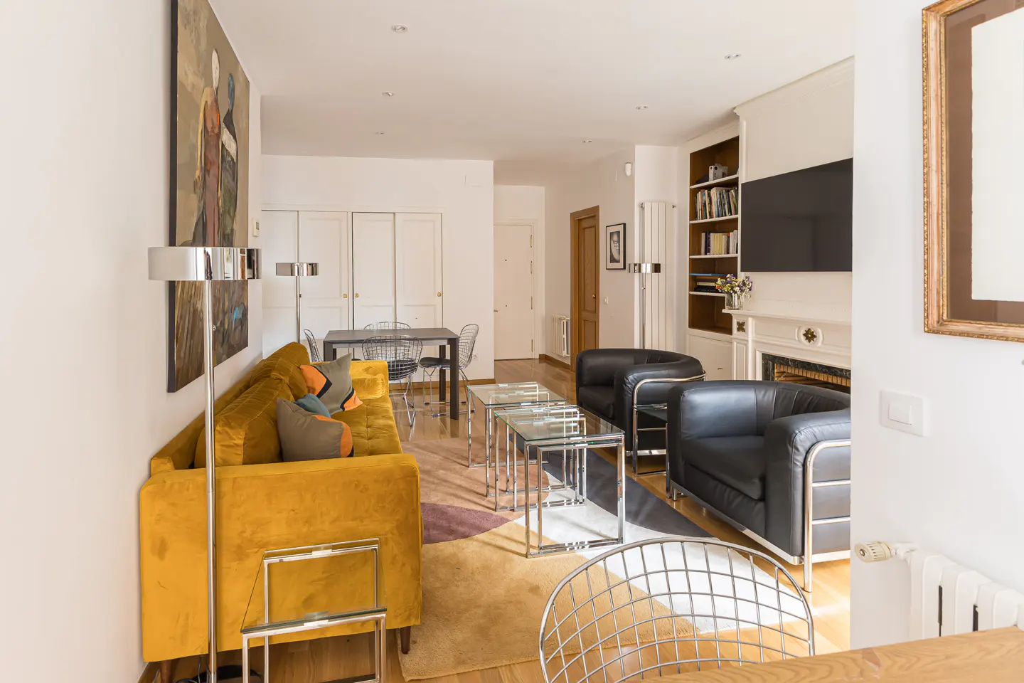Bright living room with yellow sofa, black chairs, glass tables, and a dining area. Artwork and a fireplace add character.