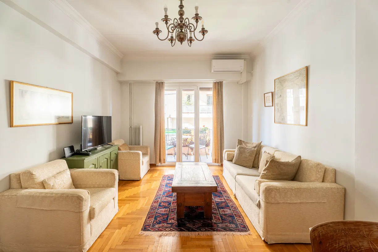 Bright living room with beige sofas, wooden floors, and a patterned rug. A TV sits on a green cabinet. French doors lead to a balcony.