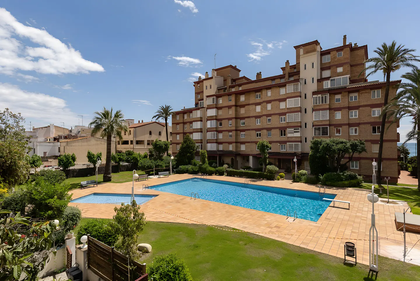 Apartment complex with a pool. The building is brick with white trim. Palm trees and green grass surround the pool. Blue sky.