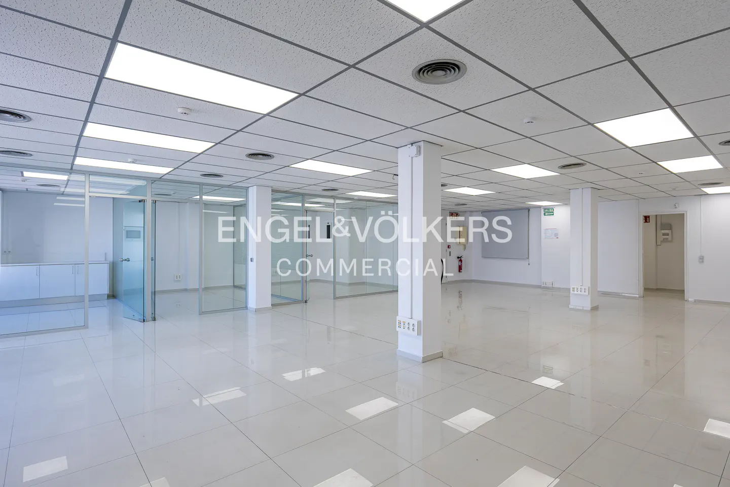 Bright, empty commercial space with white tile floors, glass-walled offices, and Engel & Völkers logo on a support column.