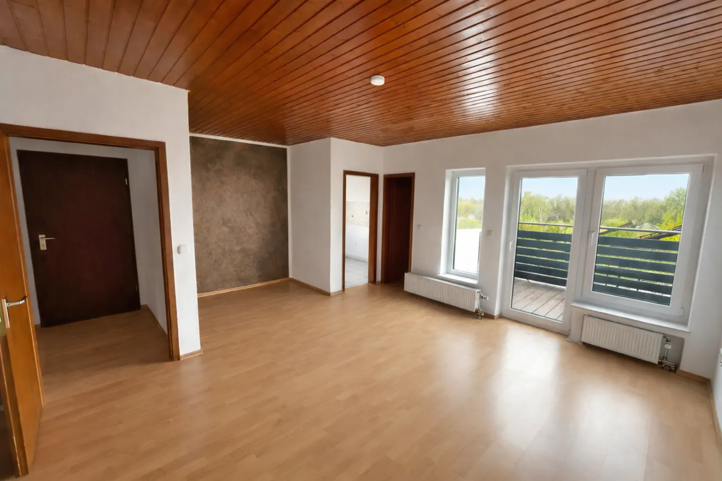 Bright, empty room with wood floors, white walls, and a wood-paneled ceiling. Windows lead to a balcony with a view of trees.