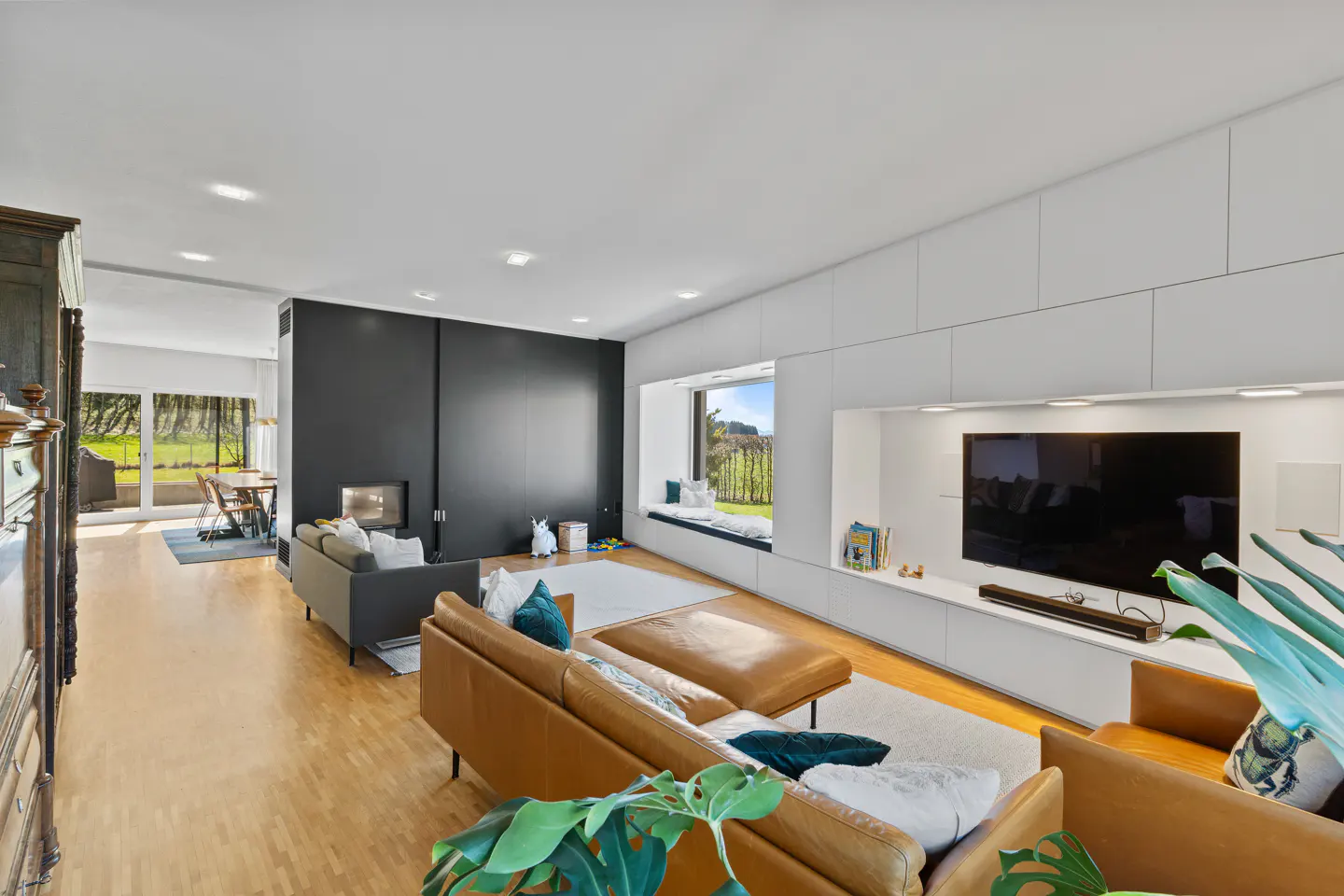 Bright living room with hardwood floors, a brown leather sofa, and a black accent wall with a fireplace. A window seat overlooks a green landscape.