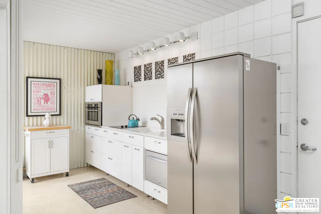 Bright kitchen with white cabinets, stainless steel fridge, oven, and dishwasher. A rolling island sits near a corrugated metal wall with framed art.