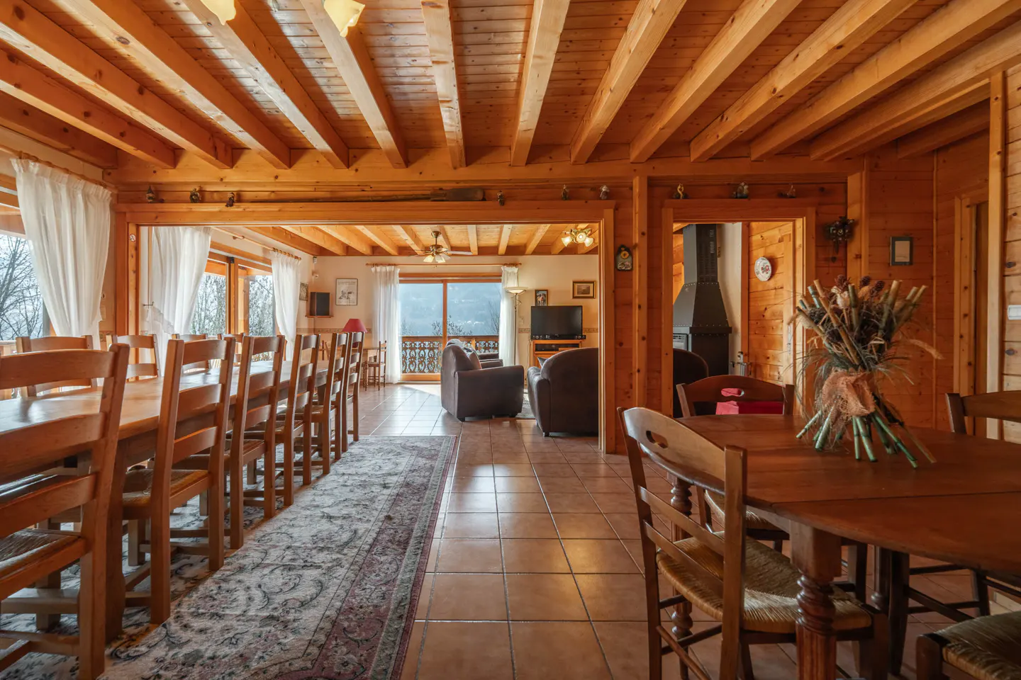 A rustic interior with wood beams, a long dining table with chairs, and a living area with brown armchairs.