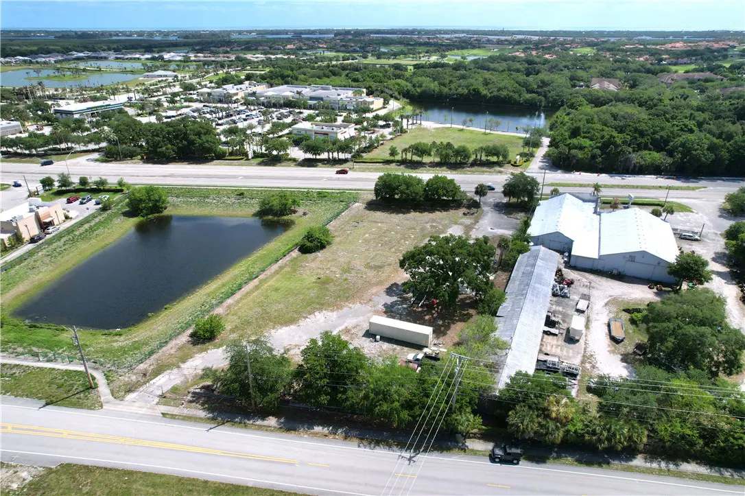 Aerial view of a commercial property with a pond, grassy area, and white buildings near a road with trees and other buildings in the background.