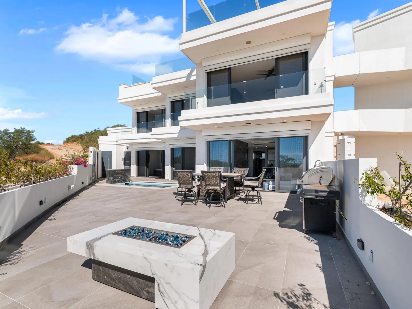 Outdoor patio of a modern white house with a fire pit, grill, table and chairs.