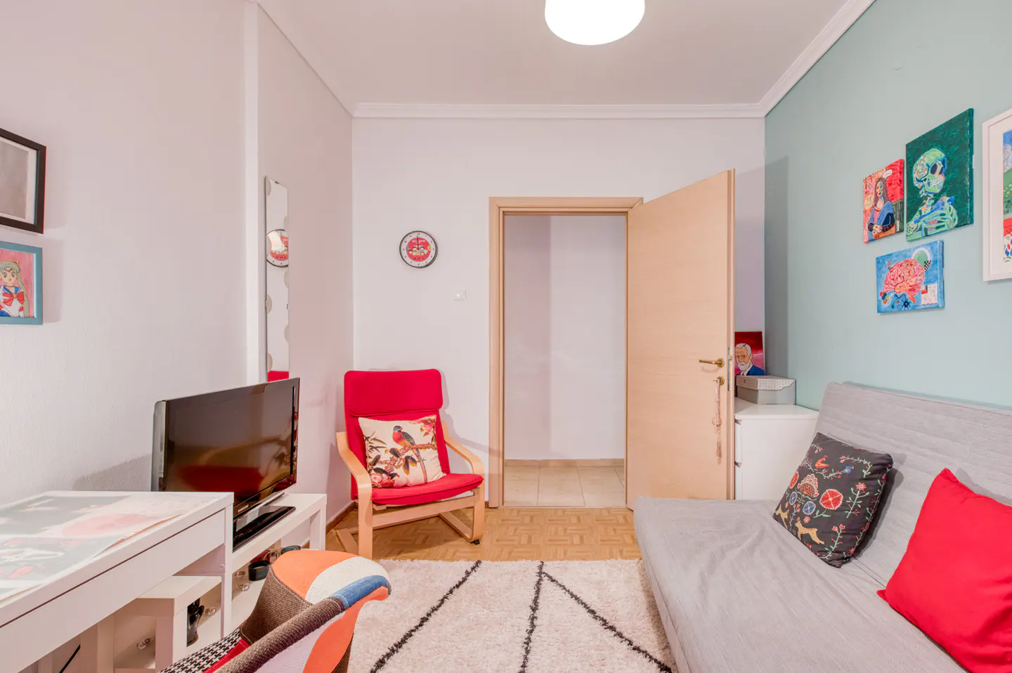 Cozy room with a gray futon, red chair, and white desk. Artwork adorns the light blue wall. A doorway leads to another room.
