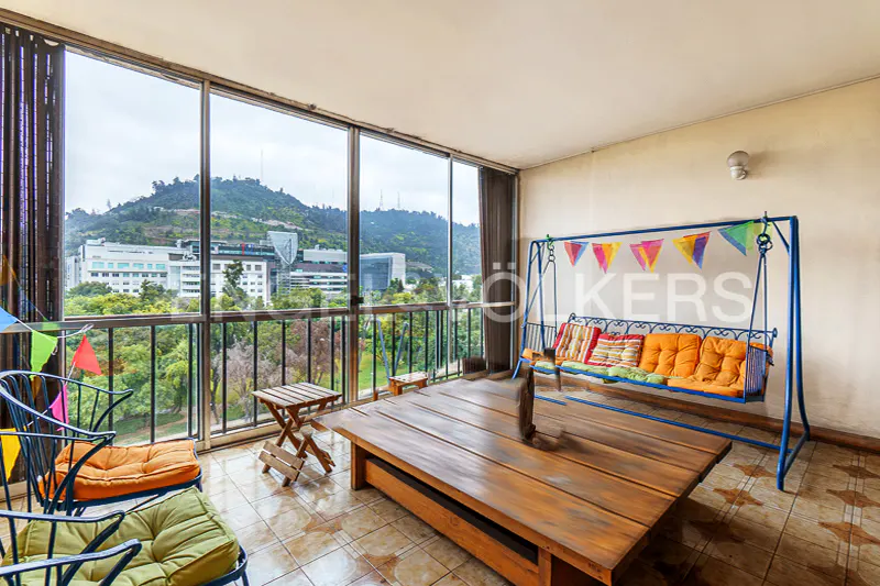 Balcony with a swing, chairs, and a large wooden table. Outside, there is a view of a city and a mountain.