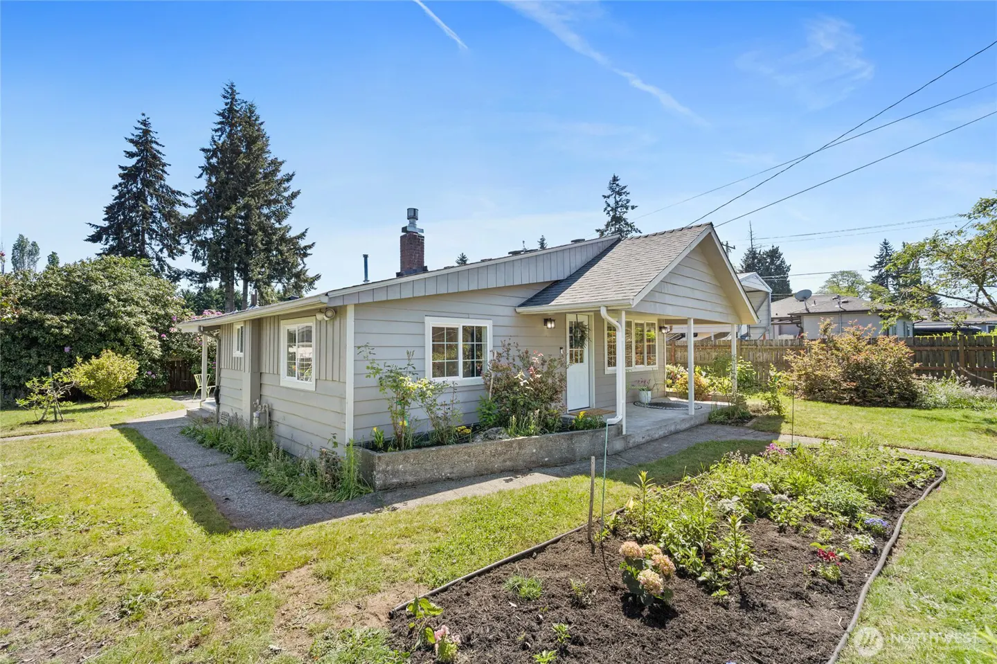 Exterior view of a one-story, light gray house with a white door, porch, and a chimney. The house has a well-maintained lawn and garden.