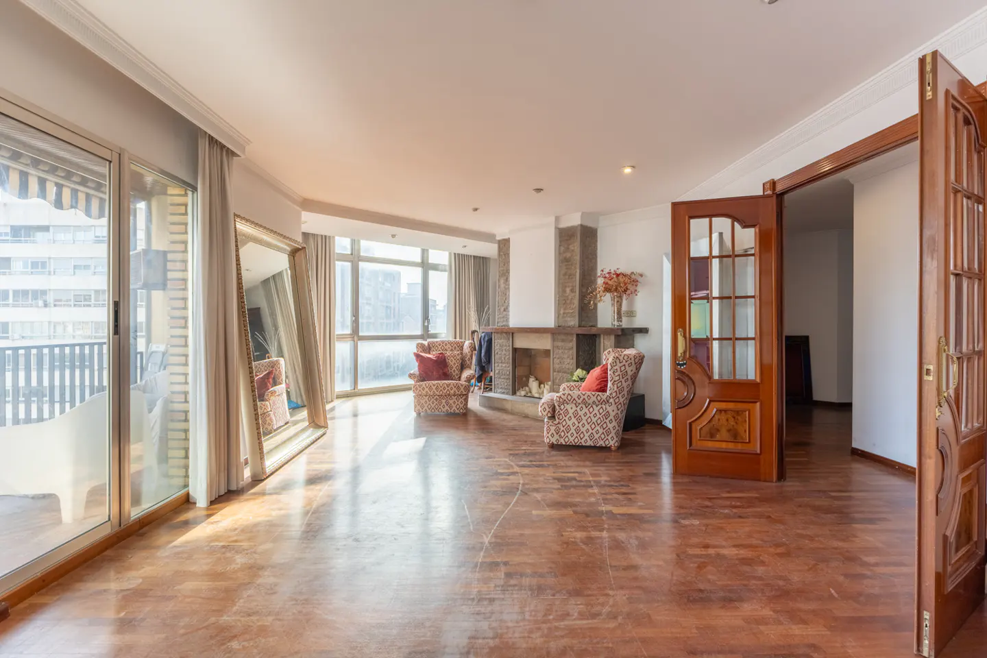 Spacious living room with hardwood floors, fireplace, and city views. Two patterned armchairs face the fireplace. A large mirror reflects the room.