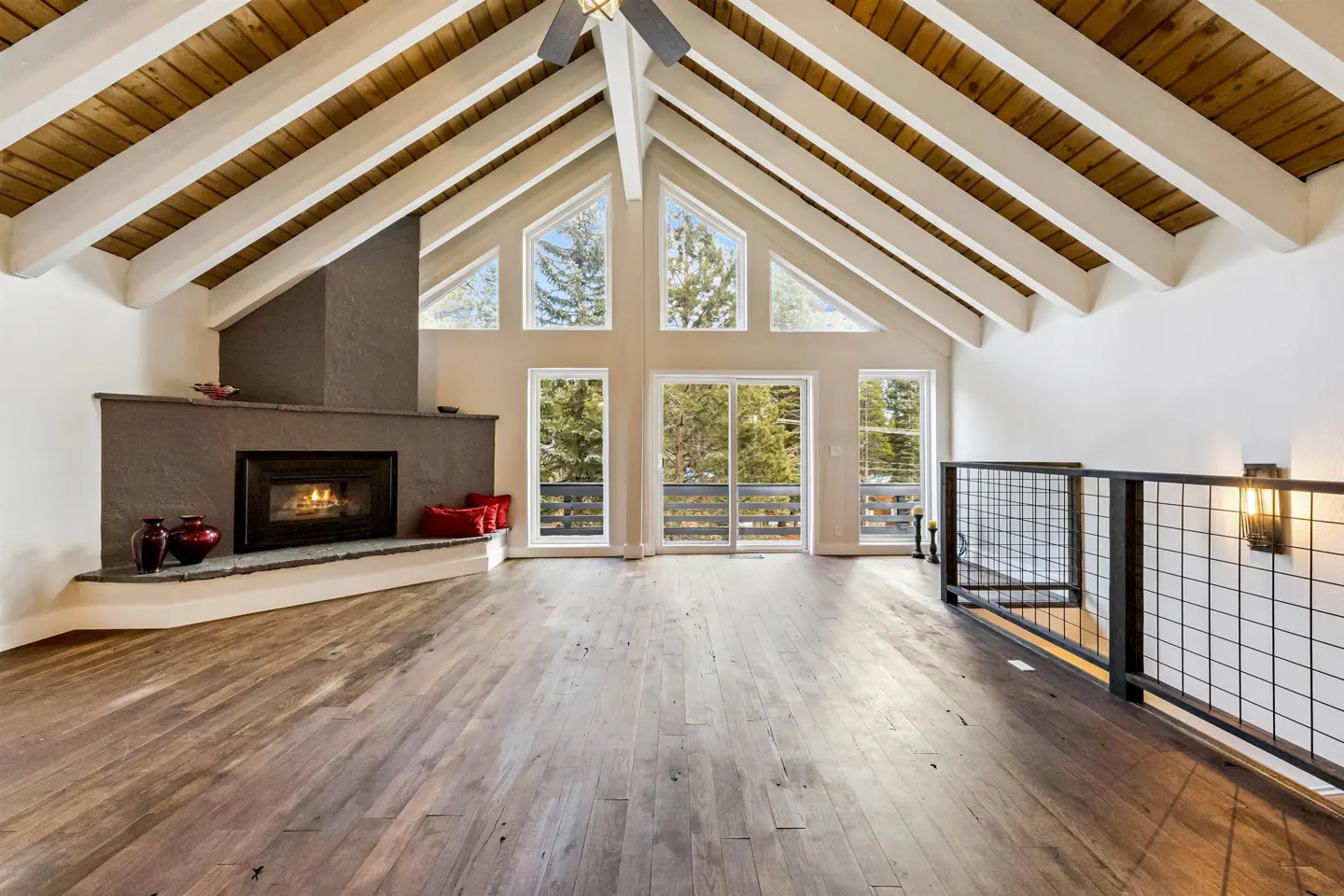 Bright living room with wood floors, a stone fireplace, and large windows overlooking trees. The ceiling has exposed wood beams.