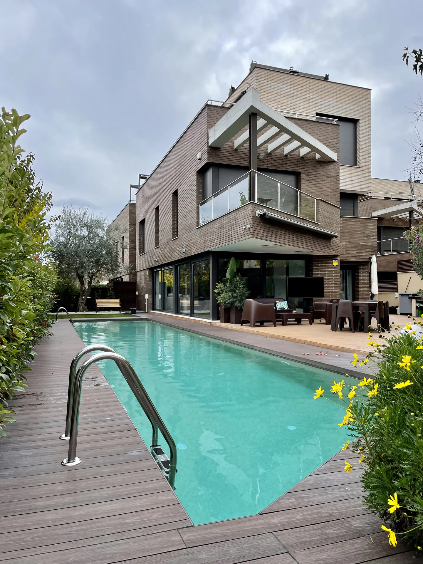 Modern brick house with a long, turquoise swimming pool. Wooden deck and yellow flowers in the foreground. Patio furniture visible.
