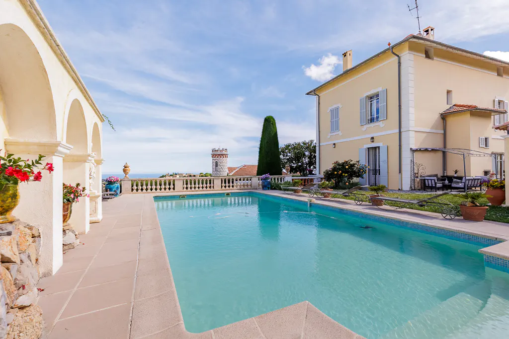 Outdoor pool with blue water, stone patio, and arched colonnade. A yellow house and tower are in the background under a blue sky.