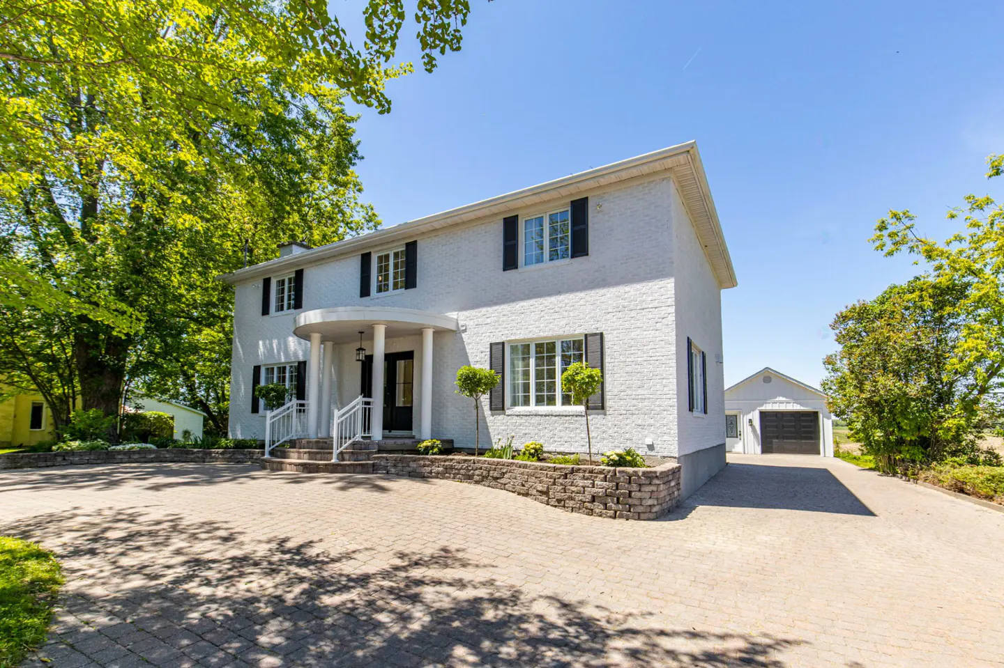 Two-story white brick house with black shutters, a curved front porch, and a detached garage on a paved driveway.