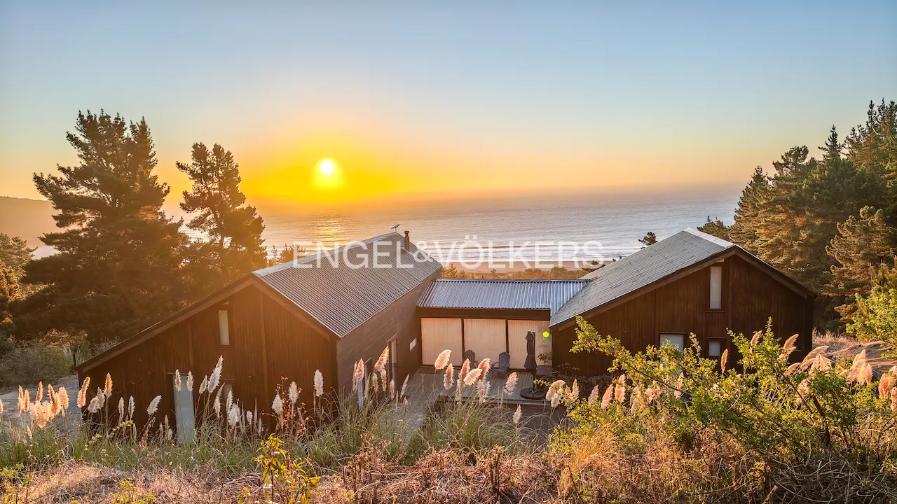 Exterior view of a modern brown house with a metal roof at sunset overlooking the ocean. Tall grass in the foreground.