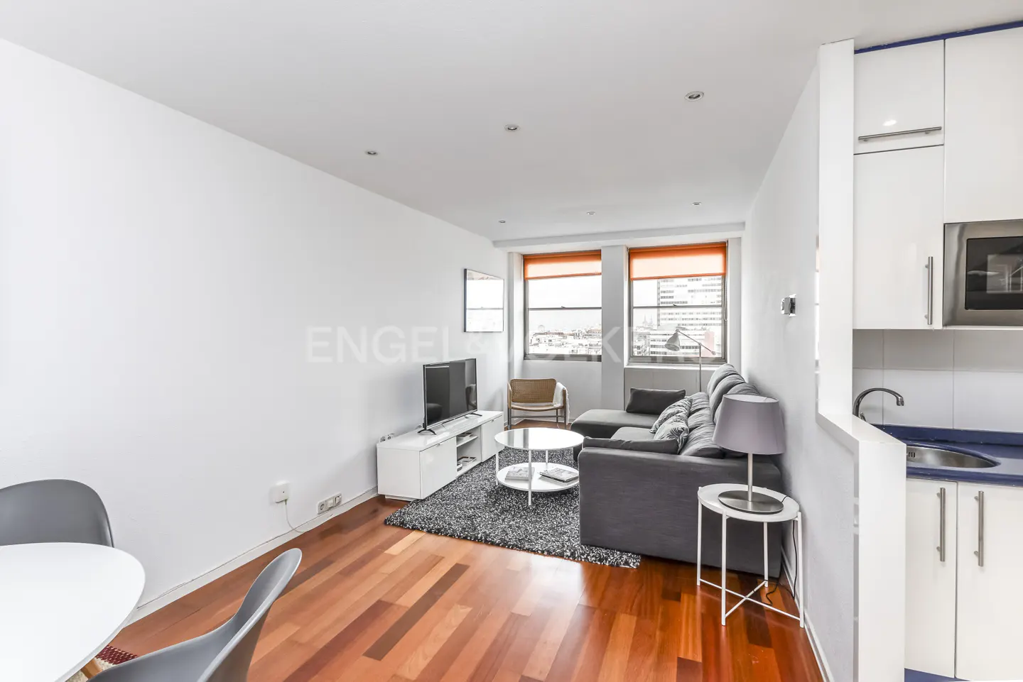 Bright apartment interior with wood floors, gray sofa, white kitchen cabinets, and city views from windows with orange blinds.