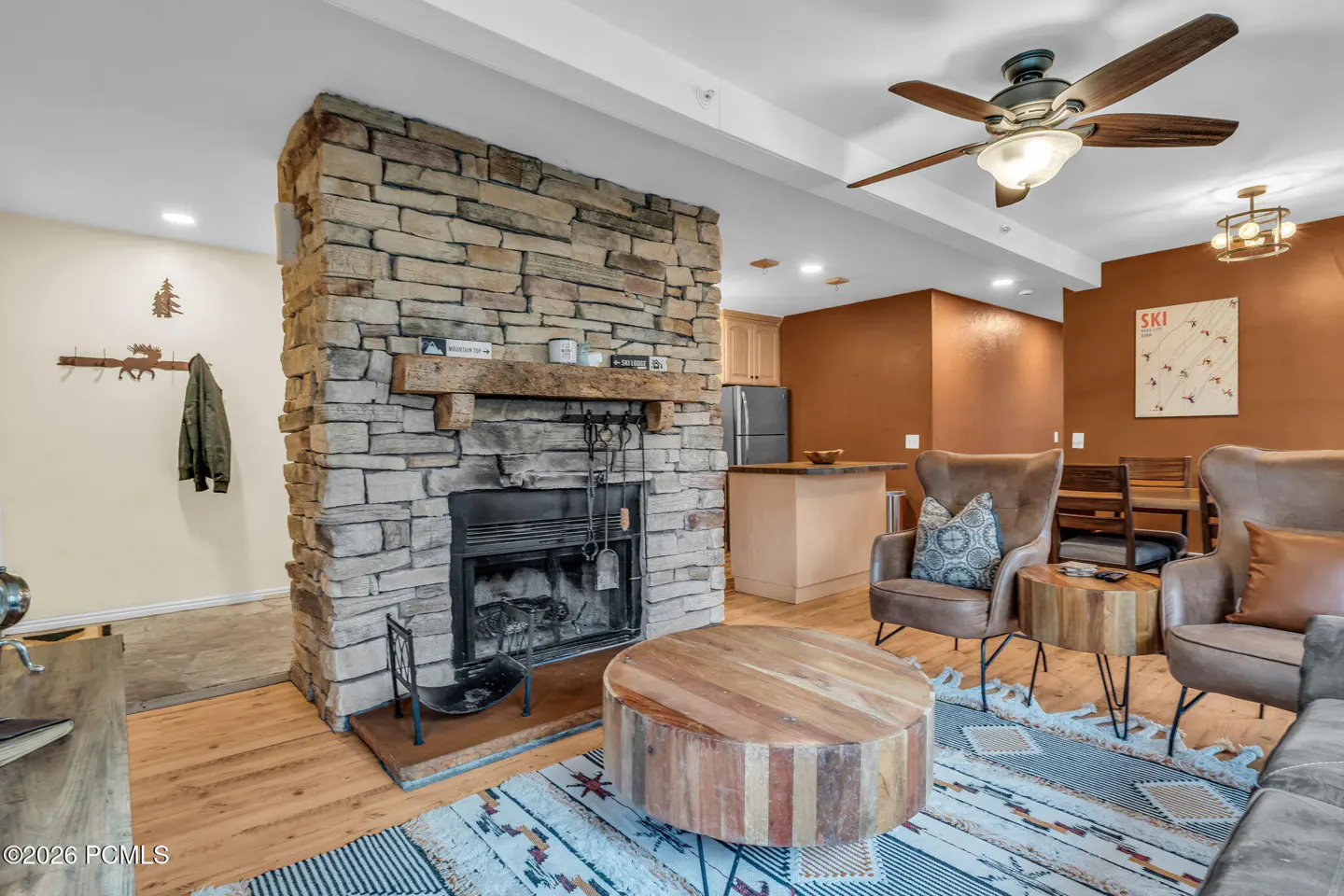Living room with a stone fireplace, wood floors, brown walls, and a round wooden coffee table. Two leather chairs face the fireplace.