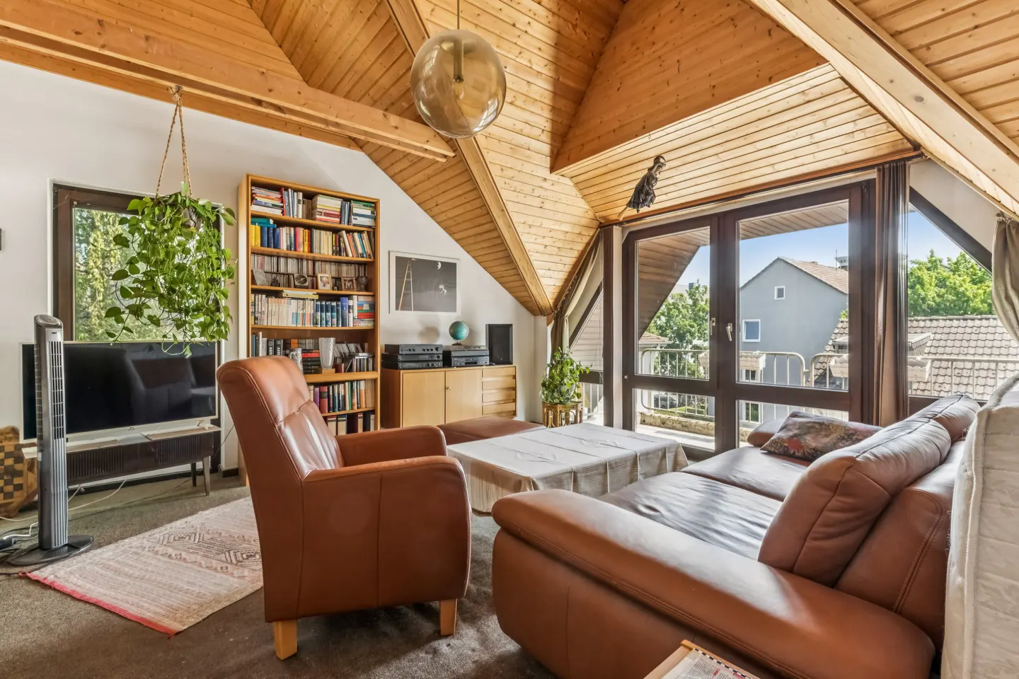 Cozy attic living room with wood-paneled vaulted ceiling, brown leather sofa and armchair, bookshelf, and balcony view.