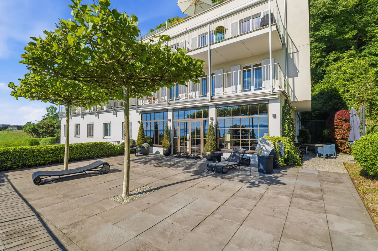 Modern white building with a stone patio, lounge chairs, and trees, set against a lush green hillside under a blue sky.