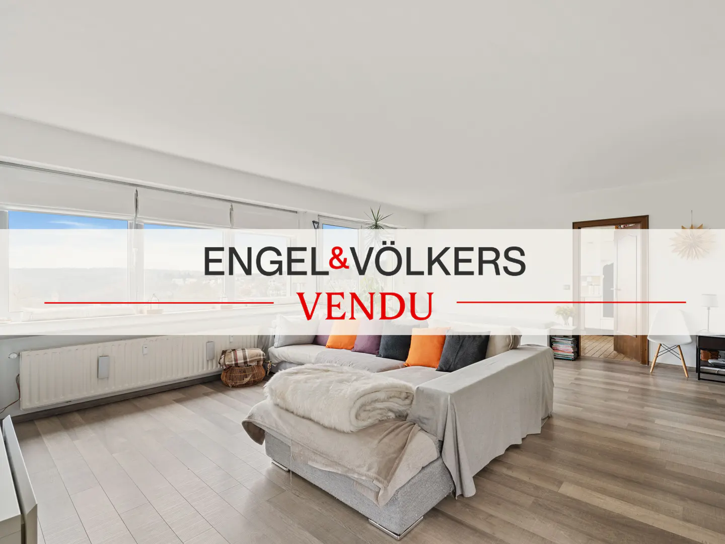 Bright living room with hardwood floors, a gray sofa with colorful pillows, and a view from the window. "Vendu" overlay.