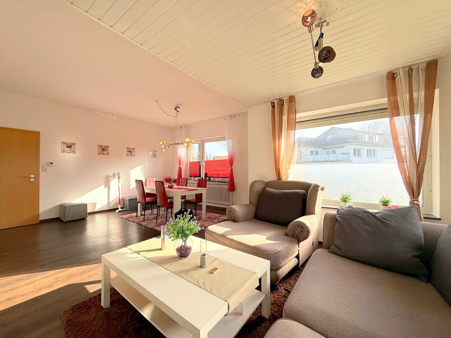 Bright living room with a white coffee table, beige sofa, and dining area with red chairs. Sunlight streams through the window.