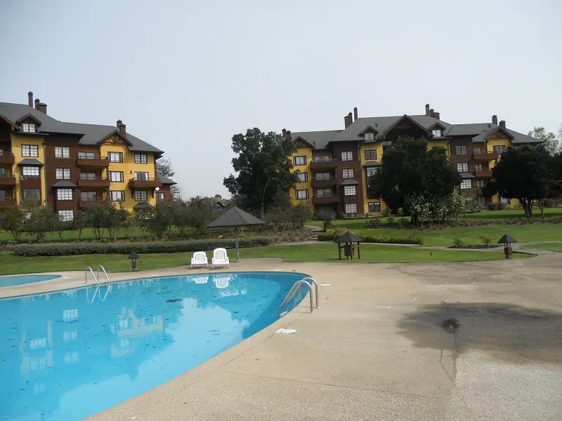 Outdoor pool with two white chairs, surrounded by green grass and trees. In the background are two yellow and brown apartment buildings.