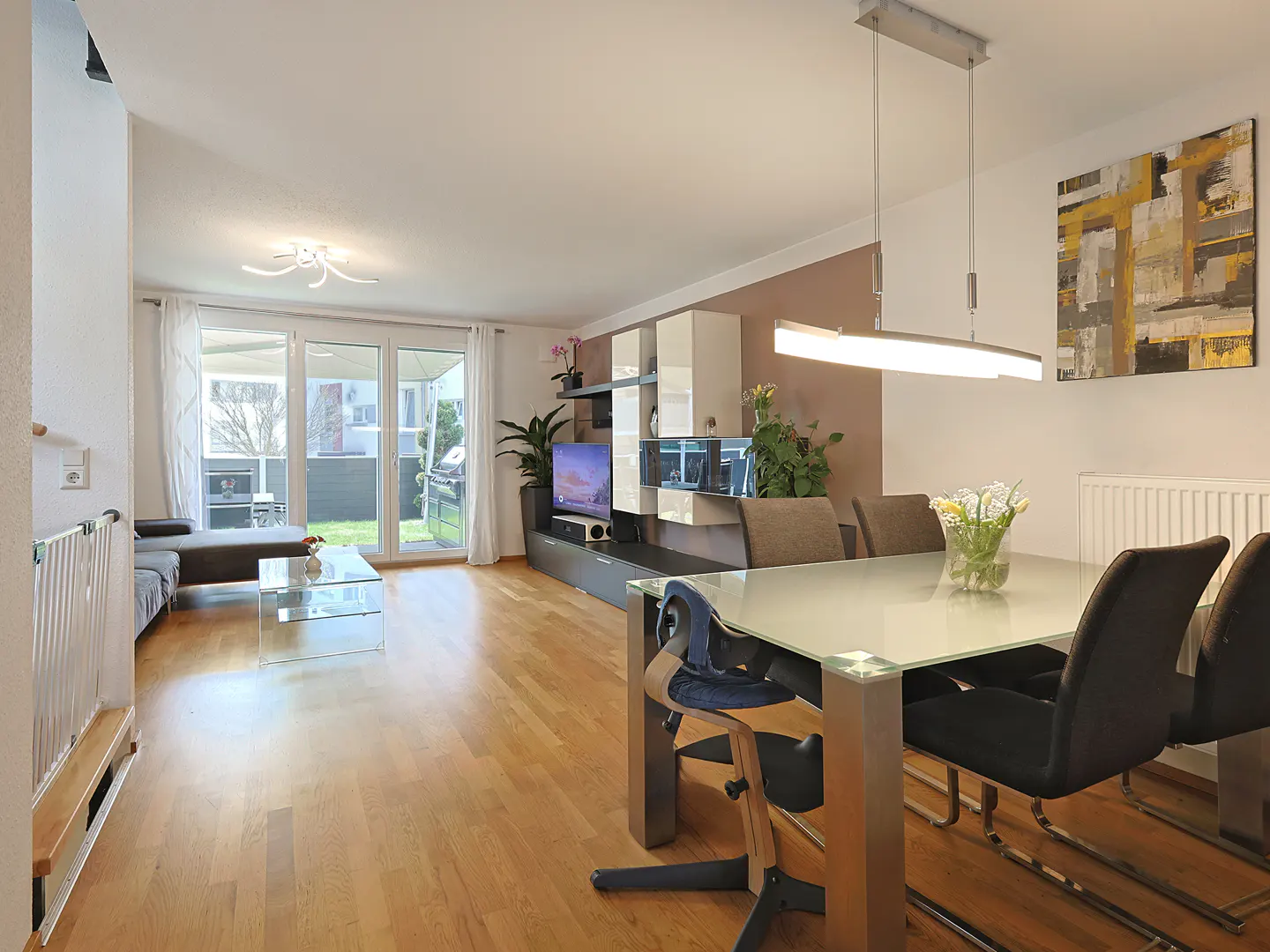 Bright, open-plan living and dining area with hardwood floors. Glass table, black chairs, and a modern light fixture. Patio doors lead to a garden.