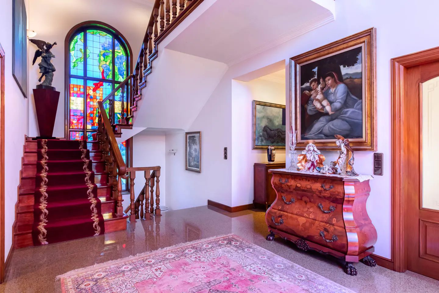 Elegant foyer with red-carpeted stairs, stained glass window, and antique wooden dresser with art.