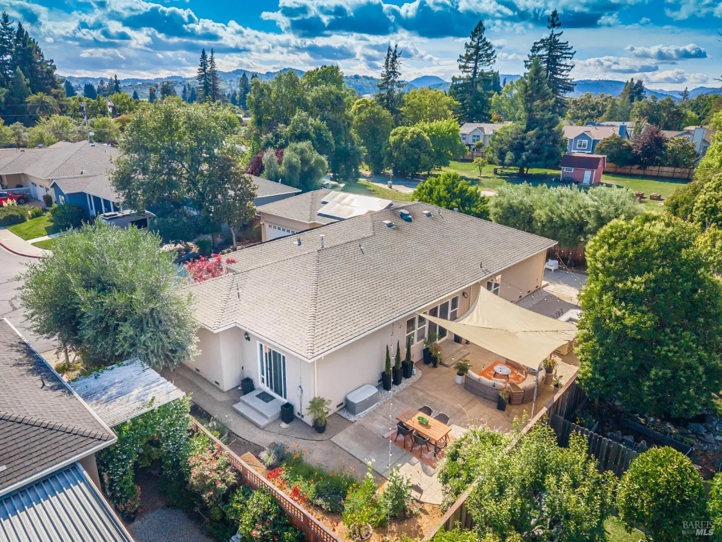 Aerial view of a beige single-story home with a patio, shade sail, outdoor dining set, and lush backyard in a suburb.