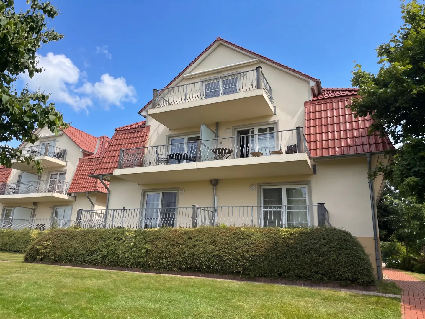 Two-story beige building with red tile roof and balconies. Green lawn and hedge in front, blue sky with clouds above.