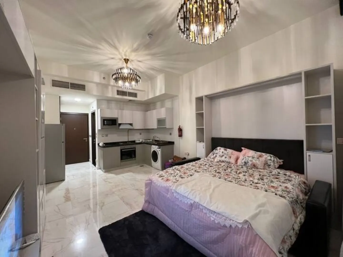 Studio apartment with a bed, kitchen, and marble floors. Two chandeliers hang from the ceiling.