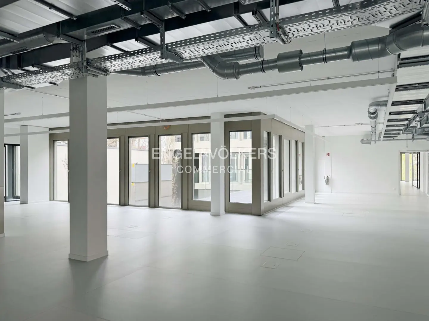 Empty commercial space with gray floors, white walls, and exposed ceiling pipes. A glass-walled office is in the center.