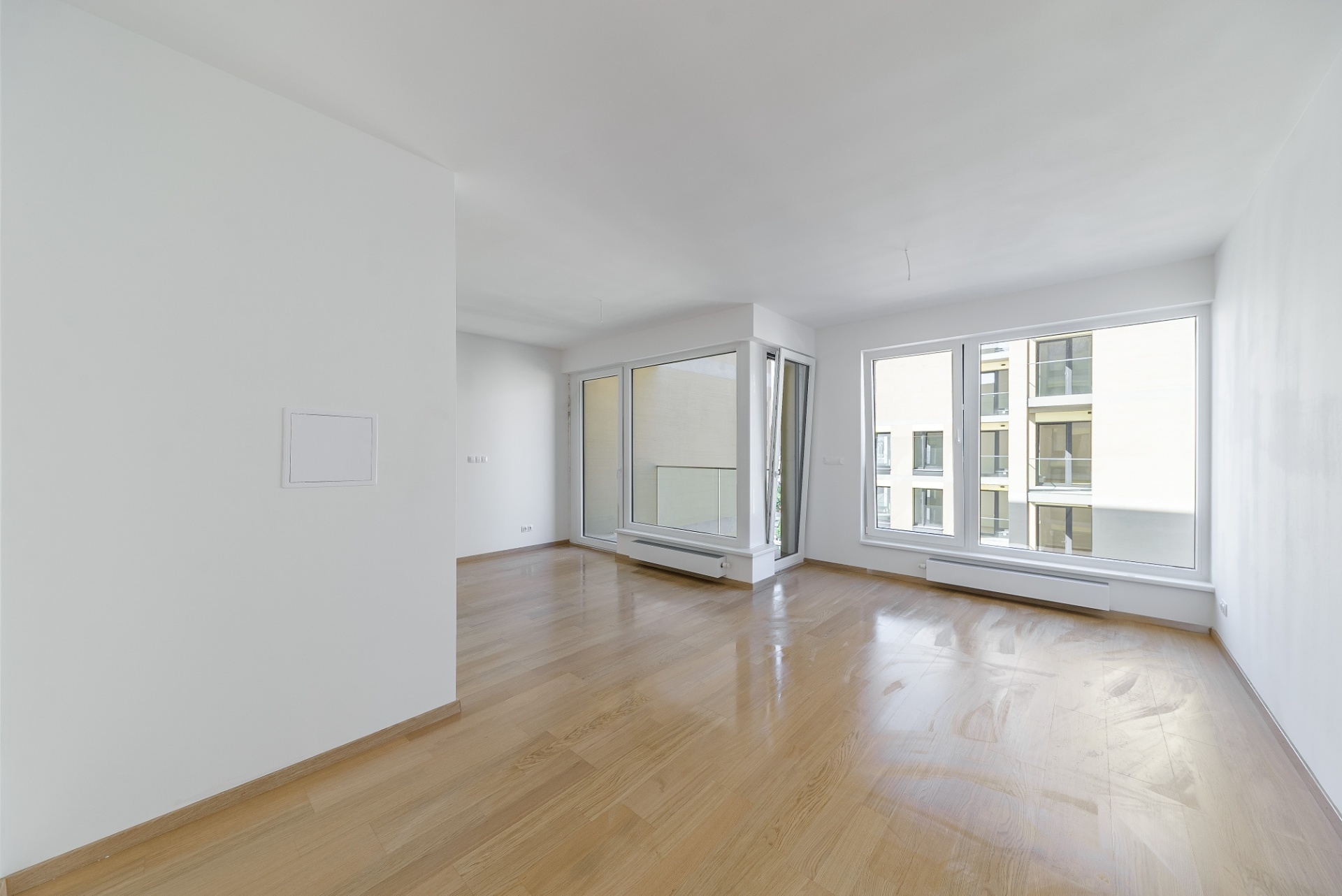 Empty, bright room with white walls, light wood flooring, and large windows overlooking a neighboring apartment building.